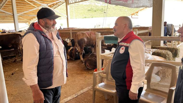 Bir kişi, diğer kişiyle sohbet ederken arka planda hayvanlar görünmektedir.