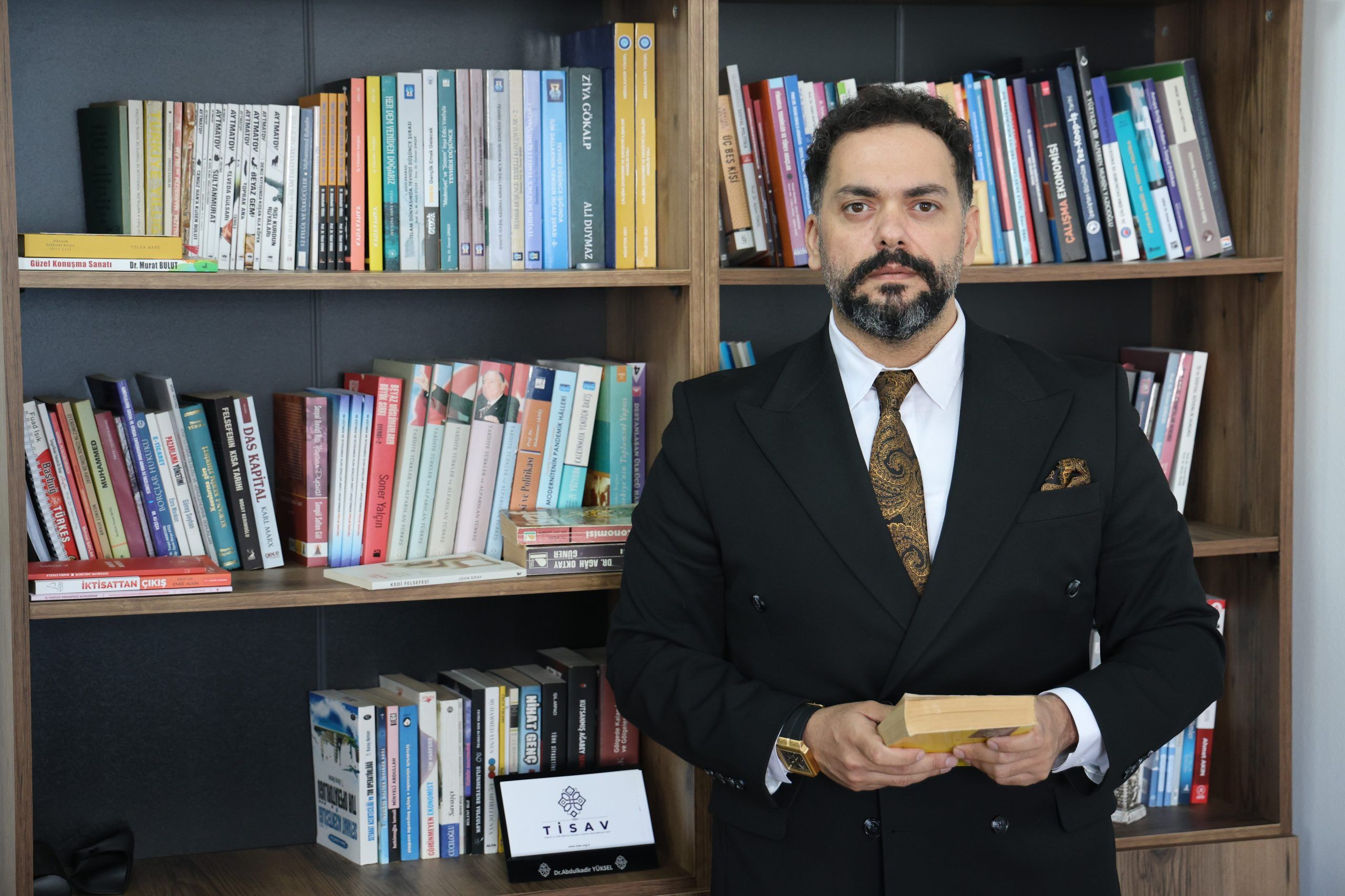 Kütüphane rafında kitapların önünde duran bir kişi, elinde bir kitap tutuyor.