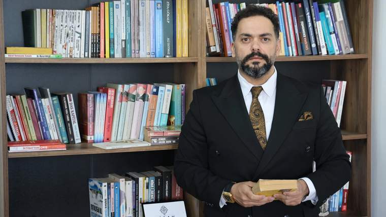 Kütüphane rafında kitapların önünde duran bir kişi, elinde bir kitap tutuyor.