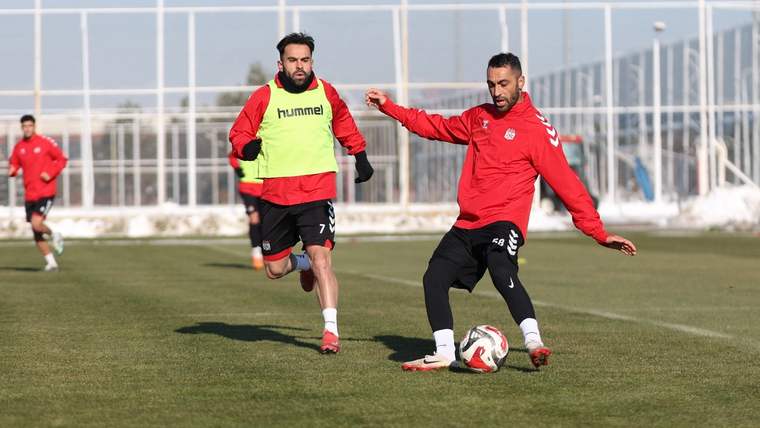 Antrenman sahasında, kırmızı forma giymiş iki futbolcu, topa vuruyor ve koşuyor.