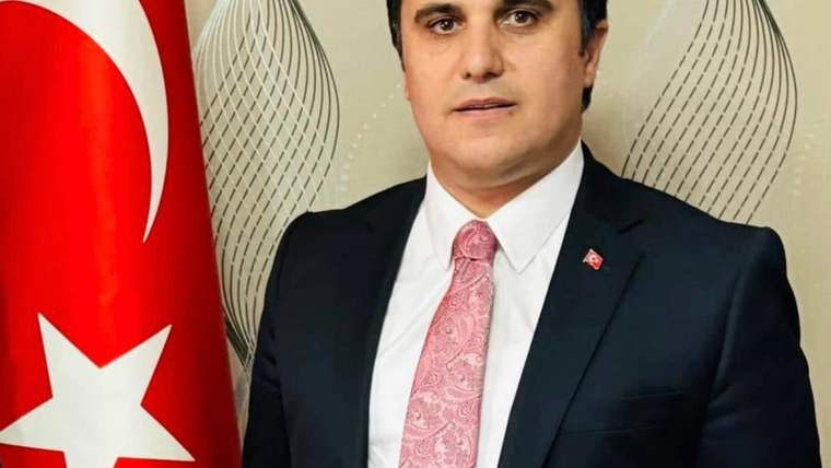Türk bayrağının yanında takım elbiseli bir erkek, desenli arka plan önünde kameraya bakıyor.