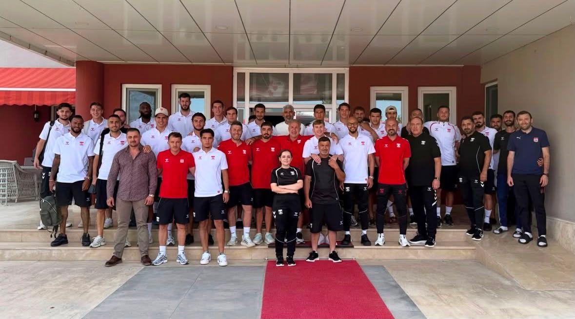 Bir bina girişinde, kırmızı halı üzerinde ve çevresinde duran spor giyimli kalabalık grup poz veriyor.