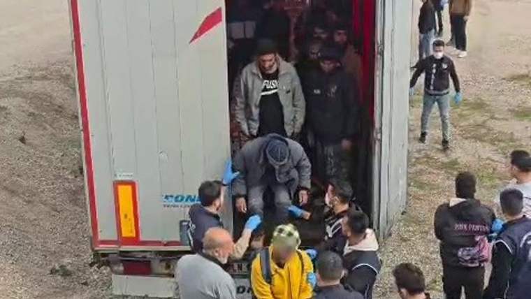 Bir tır dorsesinden inen düzensiz göçmenler, çevresinde polis ve diğer kişilerle birlikte duruyor.