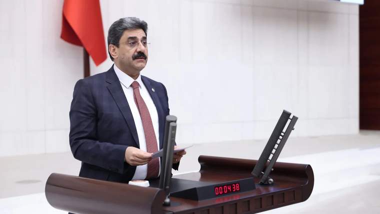 Kürsüde konuşan bir kişi, önünde bir saat ve mikrofon var.
