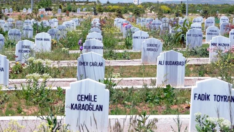 Beyaz mermer mezar taşlarının bulunduğu bir mezarlık alanı, etrafında çiçekler ve yeşil bitkiler var.