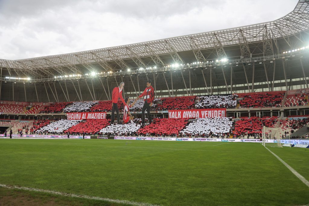 Futbol stadında yeşil saha, kırmızı-beyaz tribünler ve koreografi yapan taraftarlar görülüyor.