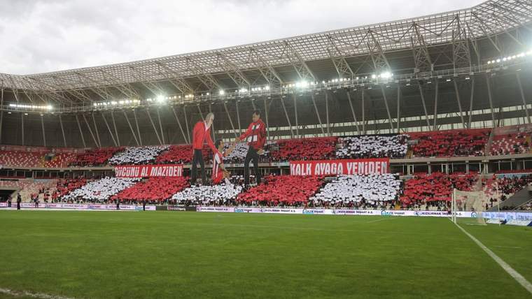 Futbol stadında yeşil saha, kırmızı-beyaz tribünler ve koreografi yapan taraftarlar görülüyor.