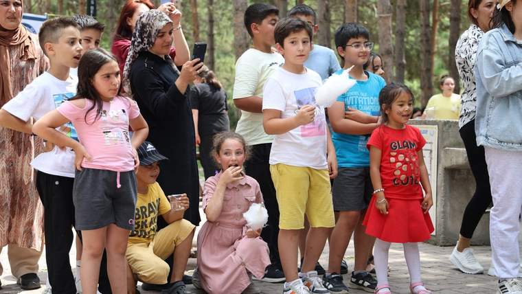 Ağaçlık alanda çocuklar ve yetişkinler bir arada duruyor; bazı çocukların elinde pamuk şeker bulunuyor.