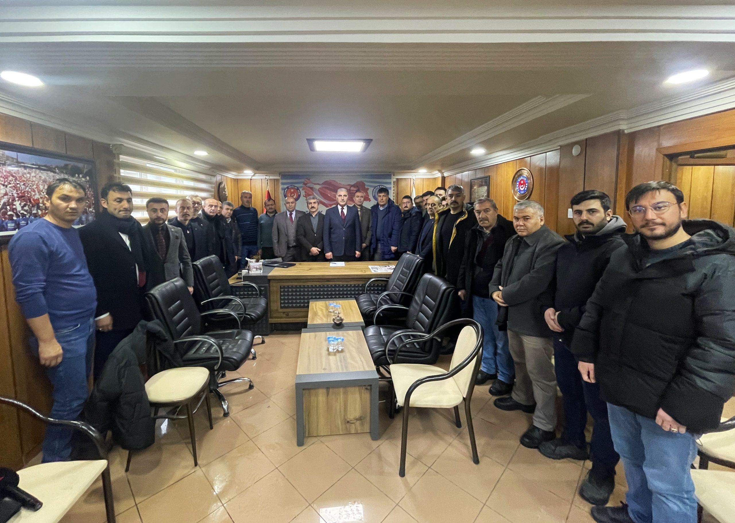 Ahşap panelli bir salonda, masa ve sandalyelerin çevresinde duran kalabalık bir grup insan görülüyor.