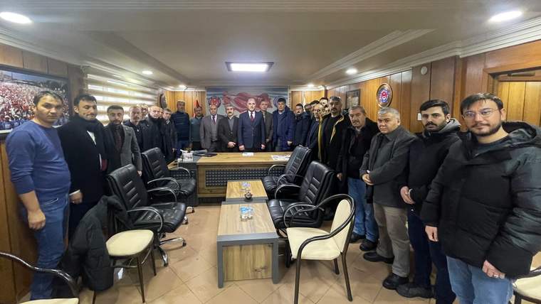 Ahşap panelli bir salonda, masa ve sandalyelerin çevresinde duran kalabalık bir grup insan görülüyor.