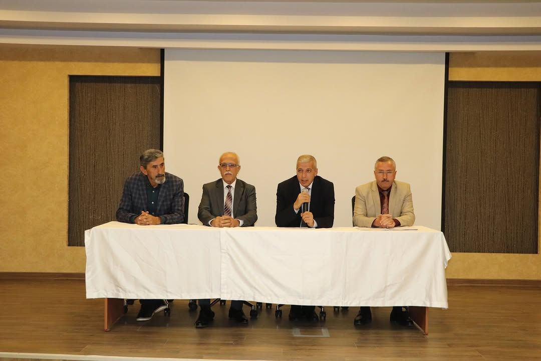 Dört kişi, beyaz örtülü bir masanın arkasında oturuyor; biri mikrofonla konuşuyor.
