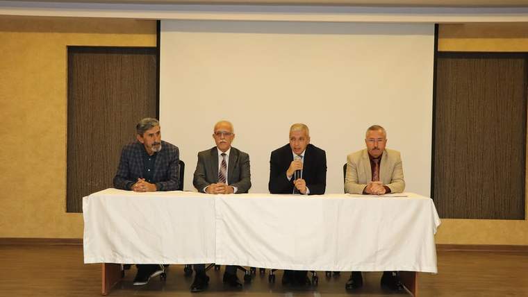 Dört kişi, beyaz örtülü bir masanın arkasında oturuyor; biri mikrofonla konuşuyor.