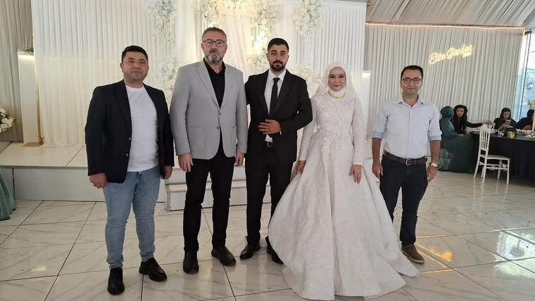 Düğün salonunda gelinlik ve takım elbise giyen çift, üç kişiyle birlikte yan yana duruyor.