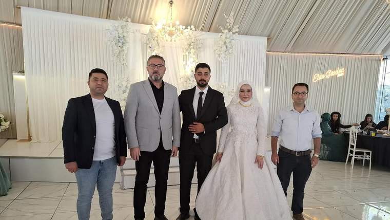 Düğün salonunda gelinlik ve takım elbise giyen çift, üç kişiyle birlikte yan yana duruyor.