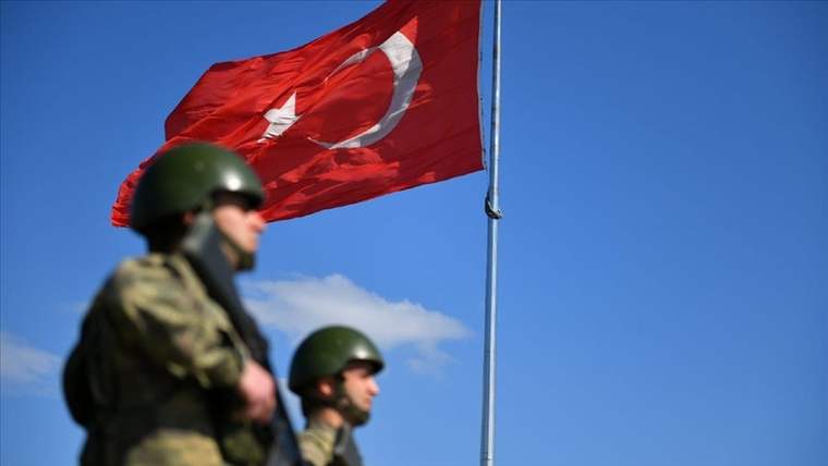 Önde iki askerin bulunduğu karede, direğe asılı Türk bayrağı rüzgârda dalgalanıyor.
