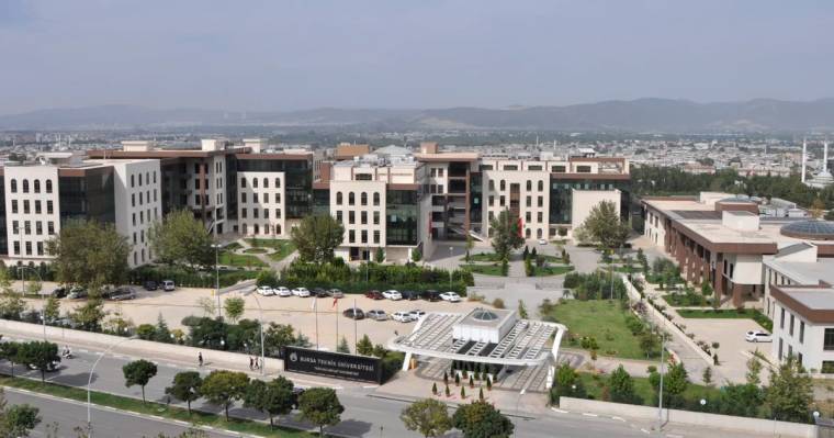 Bursa Teknik Üniversitesi yazılı tabelanın önünde, çok katlı binalar ve ağaçlı kampüs alanı görülüyor.