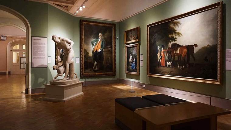 Bir sanat galerisi içindeki heykel ve tablolar, duvarlarda asılı. Zemin ahşap.