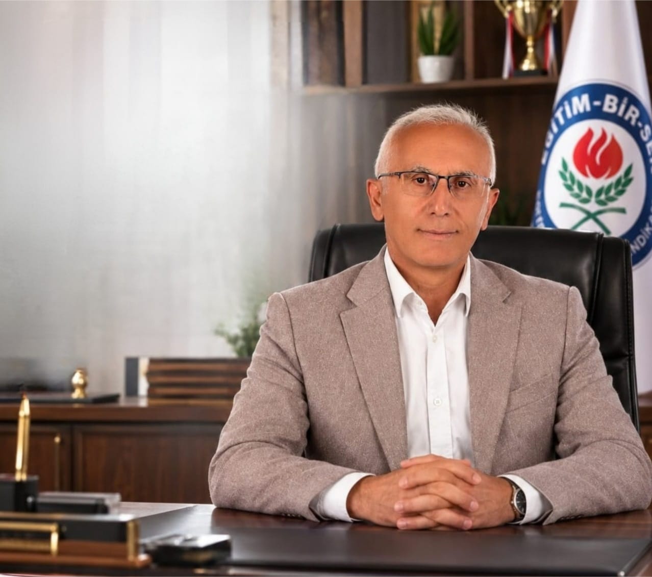 Masa başında oturan gözlüklü bir kişi, arkasındaki Eğitim-Bir-Sen logolu bayrakla kameraya bakıyor.