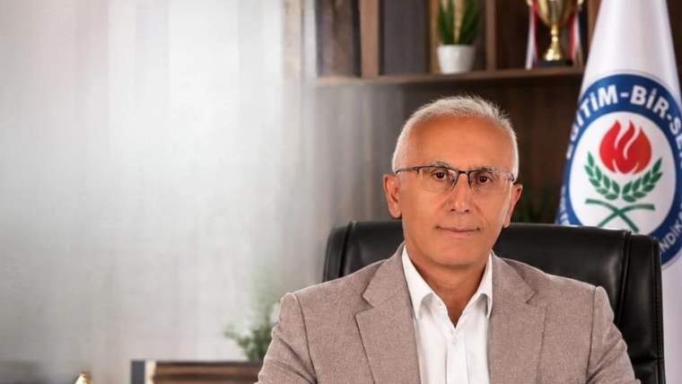 Masa başında oturan gözlüklü bir kişi, arkasındaki Eğitim-Bir-Sen logolu bayrakla kameraya bakıyor.