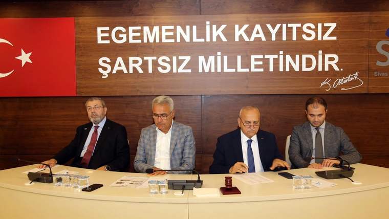 Bir toplantıda dört kişi, masanın etrafında oturuyor; arka planda Türk bayrağı ve yazı var.