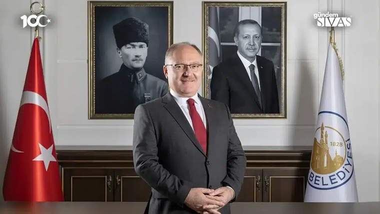 Takım elbiseli bir kişi, Türk bayrağı ve belediye flamaları arasında, iki portre önünde ayakta duruyor.