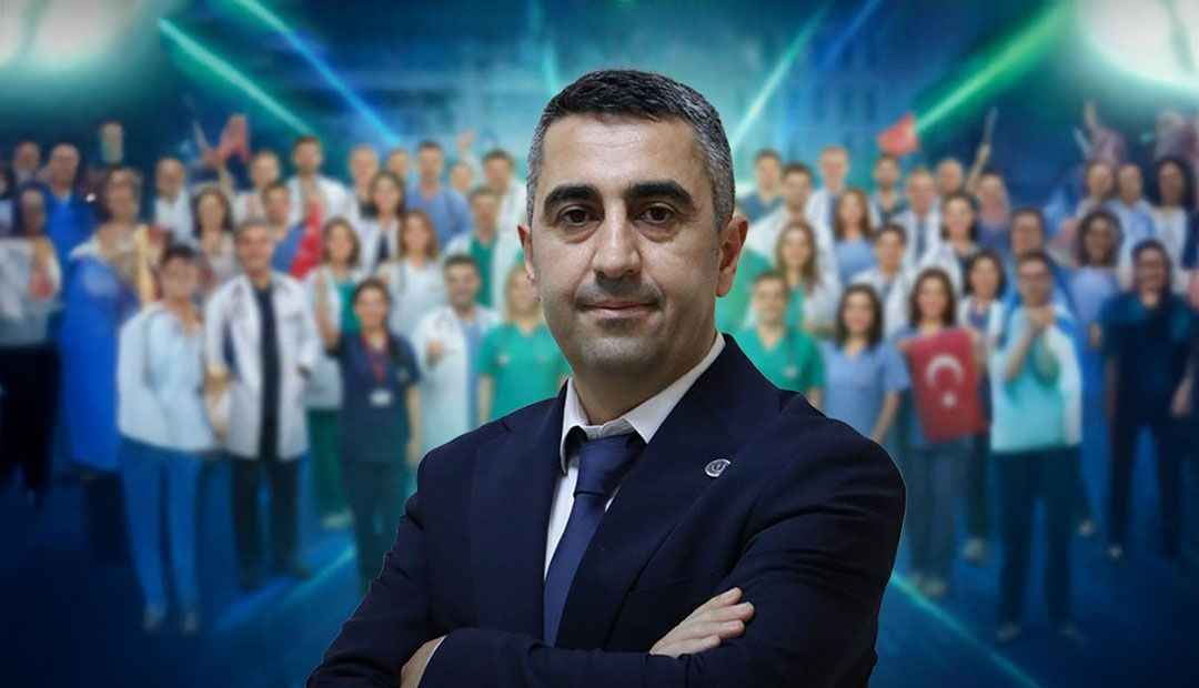 Kollarını kavuşturmuş bir kişi, arka planda sağlık çalışanlarıyla birlikte poz veriyor.