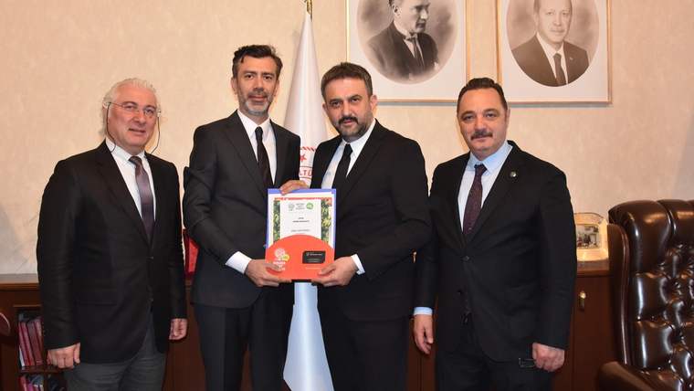 Bir odada takım elbiseli dört erkek, üzerinde belge bulunan kırmızı plaketi birlikte tutuyor; arkada bayraklar ve portreler var.