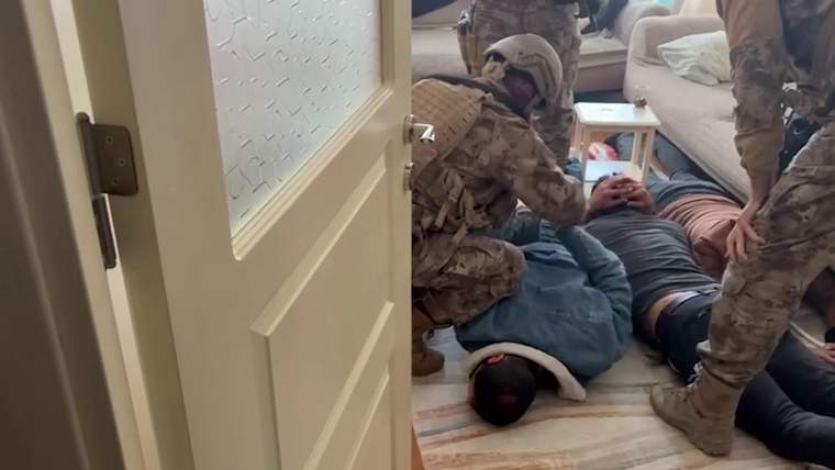Kamuflajlı görevliler, bir evin salonunda yerde yatan iki kişiyi kelepçelerken görülüyor.