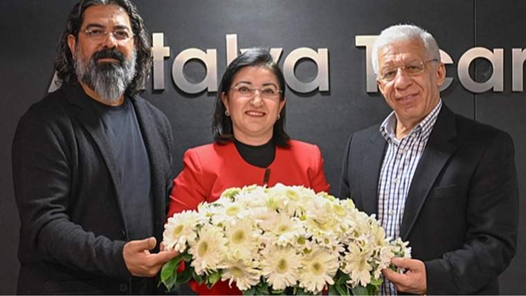 Üç kişi, siyah arka plan önünde büyük bir buket beyaz çiçek tutarak yan yana duruyor.