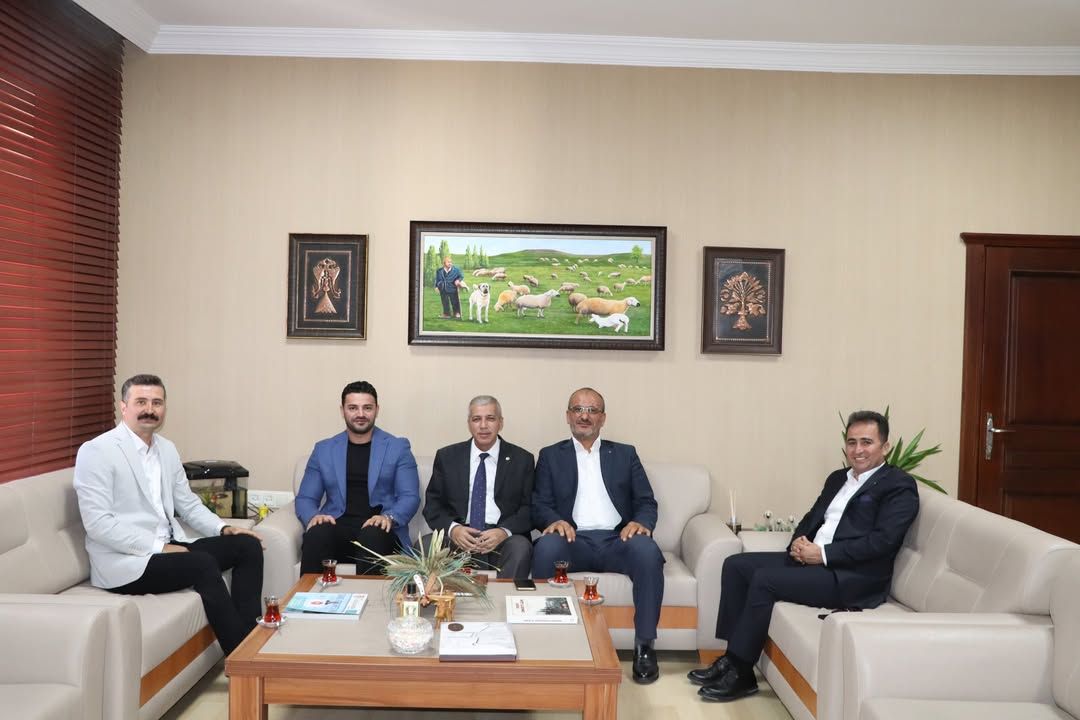Bir odada koltuklarda oturan beş kişi, orta sehpanın çevresinde poz veriyor; arkalarında tablolar bulunuyor.