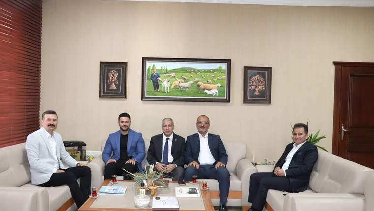 Bir odada koltuklarda oturan beş kişi, orta sehpanın çevresinde poz veriyor; arkalarında tablolar bulunuyor.