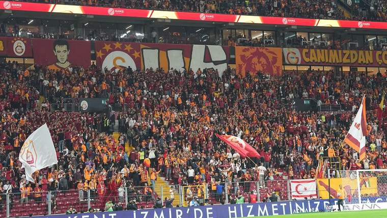 Tribünde Galatasaray renkleri ve bayraklarıyla toplanan kalabalık taraftar grubu, pankartlar arasında görülüyor.