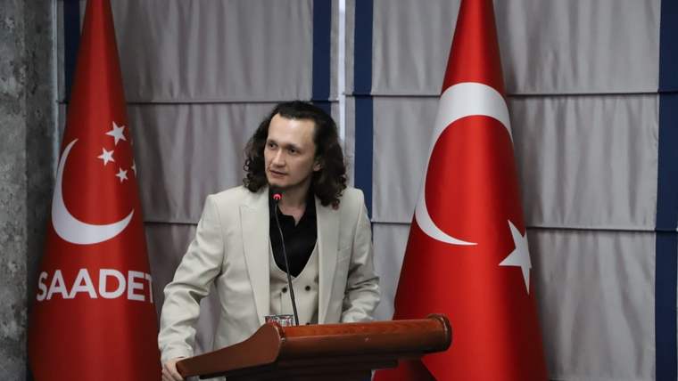Kürsüde konuşan uzun saçlı bir kişi, iki Türk bayrağı ve Saadet Partisi bayrağı önünde duruyor.