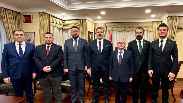 Bir odada takım elbiseli yedi erkek yan yana duruyor; duvarda çerçeveli Türk bayrağı bulunuyor.