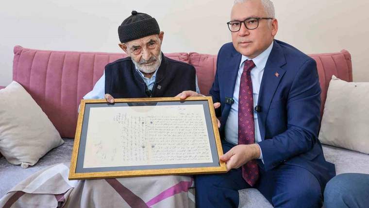 Koltuğa oturan iki kişi, üzerinde el yazısı bulunan çerçeveli bir mektubu birlikte tutuyor.