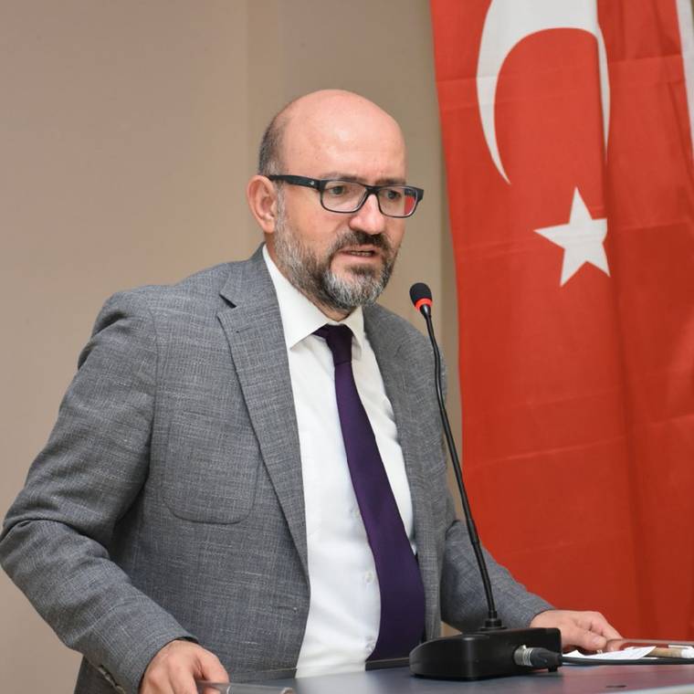 Gözlüklü ve sakallı bir kişi, mikrofon önünde konuşurken arkasında Türk bayrağı görülüyor.