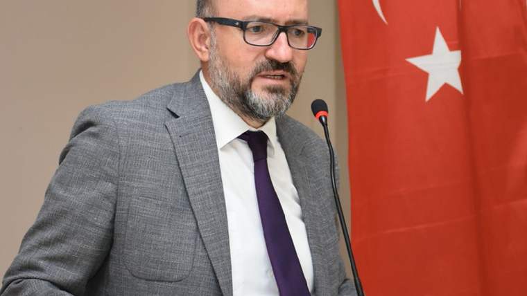 Gözlüklü ve sakallı bir kişi, mikrofon önünde konuşurken arkasında Türk bayrağı görülüyor.