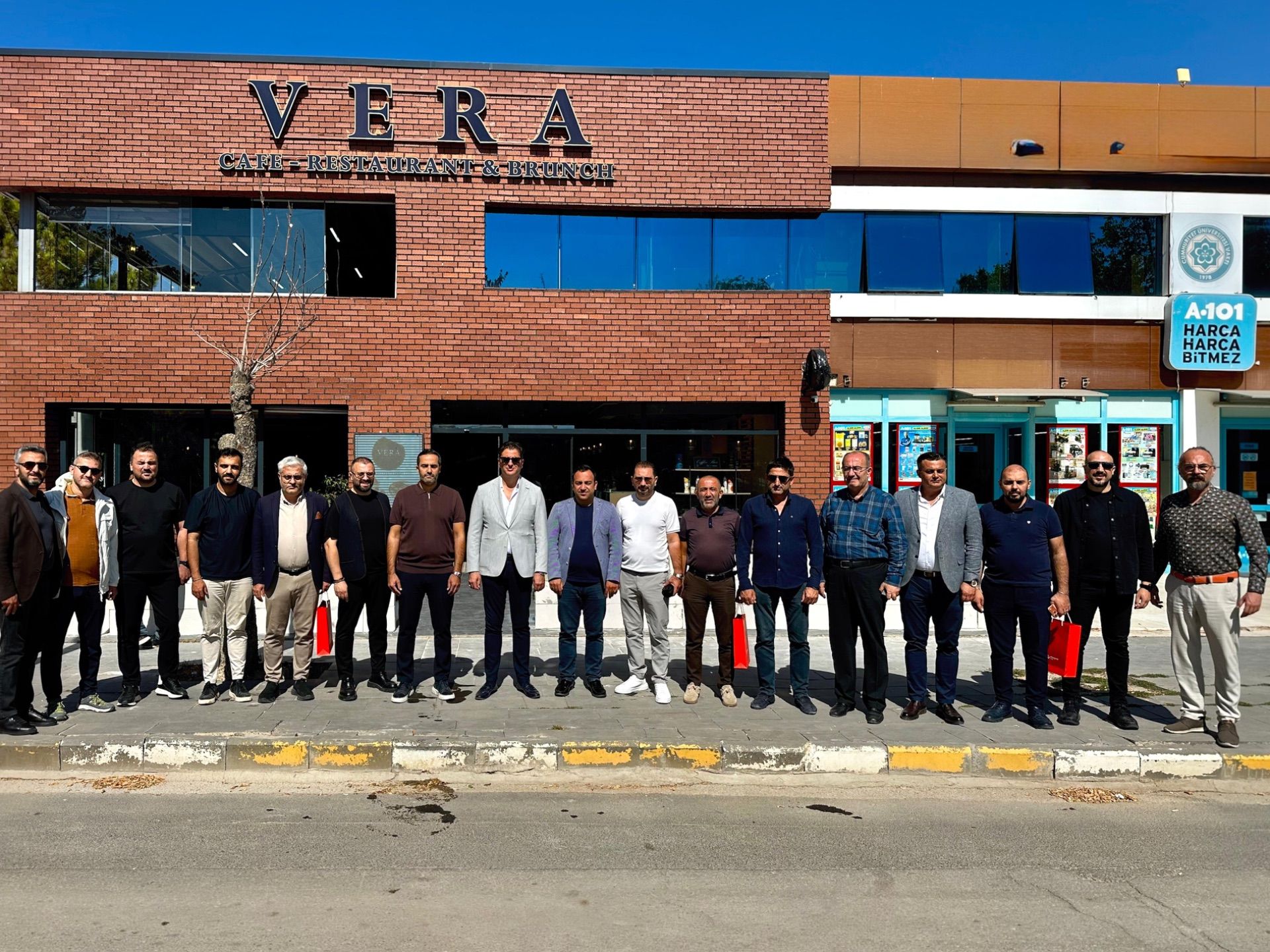Üzerinde VERA yazan binanın önünde, kaldırımda yan yana duran bir grup insan görülüyor.