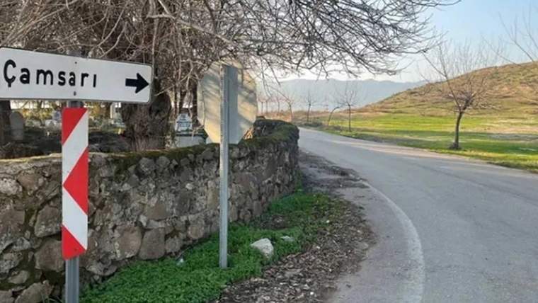 Kırsal yolda taş duvar, sağa yönü gösteren kısmen görünen tabela ve kırmızı-beyaz trafik levhası bulunuyor.