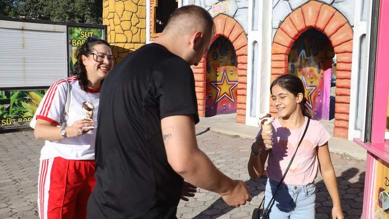 Lunaparkta, bir çift çocuklara dondurma ikram ediyor; arka planda renkli binalar görünmekte.