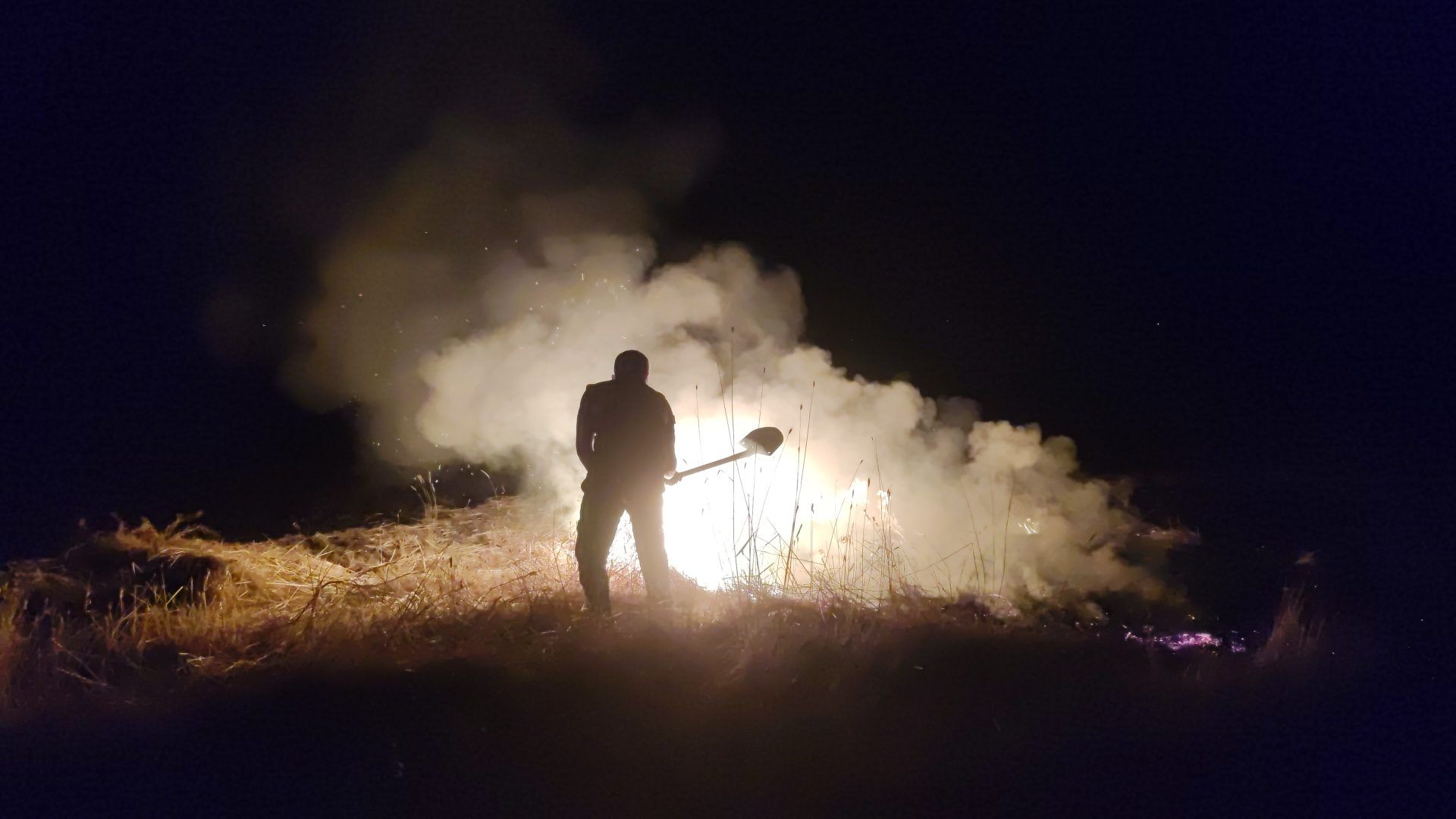 Gece karanlığında, kuru otların arasında duman ve alevlerin önünde duran bir kişinin silueti görülüyor.