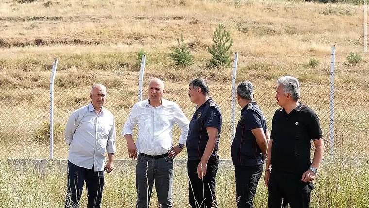 Kurumuş otlarla kaplı arazide, tel çit önünde duran beş kişi birlikte görülüyor.