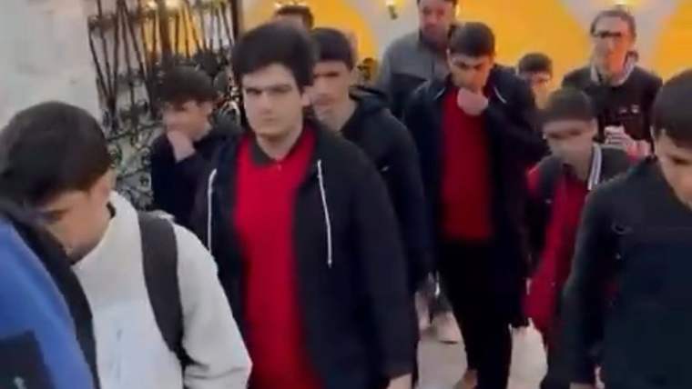 Sırt çantalı öğrenciler ve birkaç yetişkin, iç mekânda bir kapıdan geçerek ilerliyor.