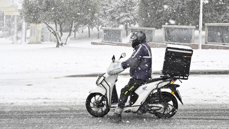 Kar yağışı altında, kasklı bir motokurye beyaz motosikletiyle karlı yolda ilerliyor.