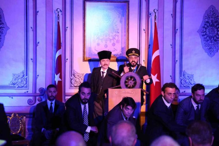 Mor ışıklarla aydınlatılan salonda, iki kişi direksiyon biçimli kürsüde dururken Türk bayrakları görülüyor.