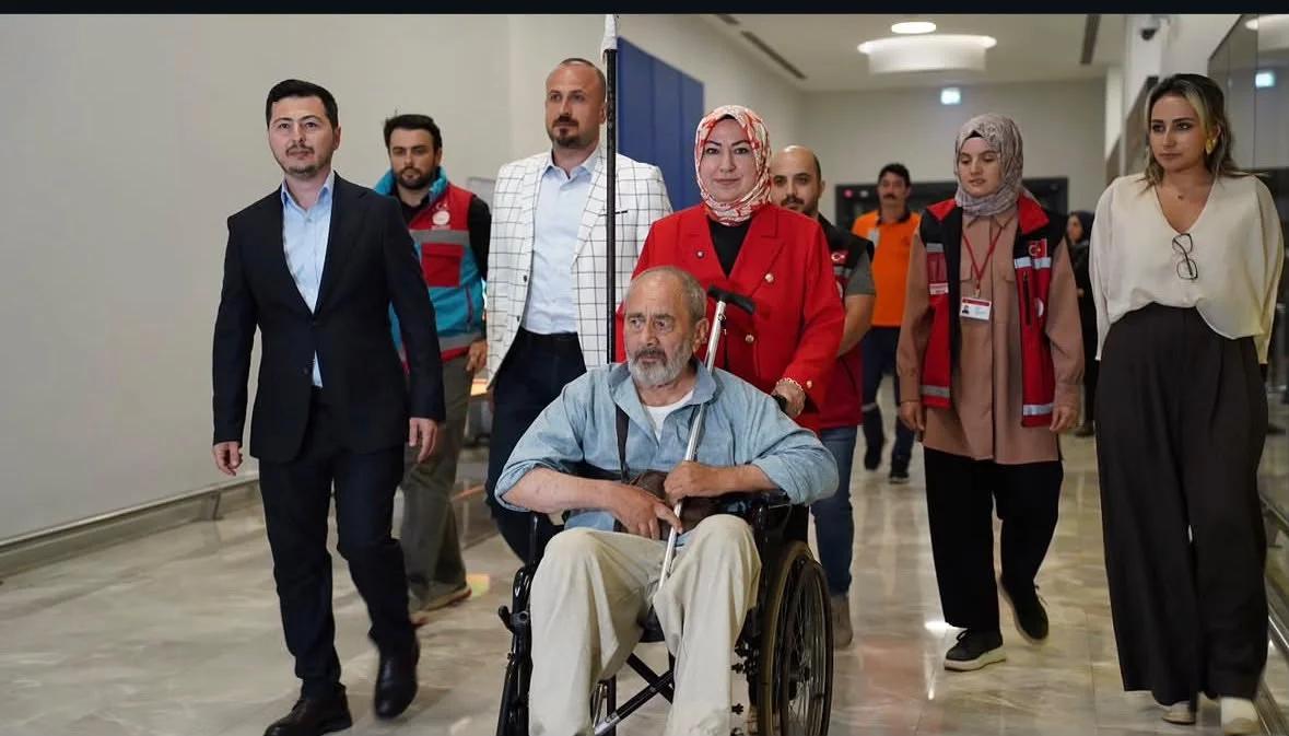 Tekerlekli sandalyede oturan bir adam, yanında yürüyen kalabalıkla birlikte bir mekandan çıkıyor.