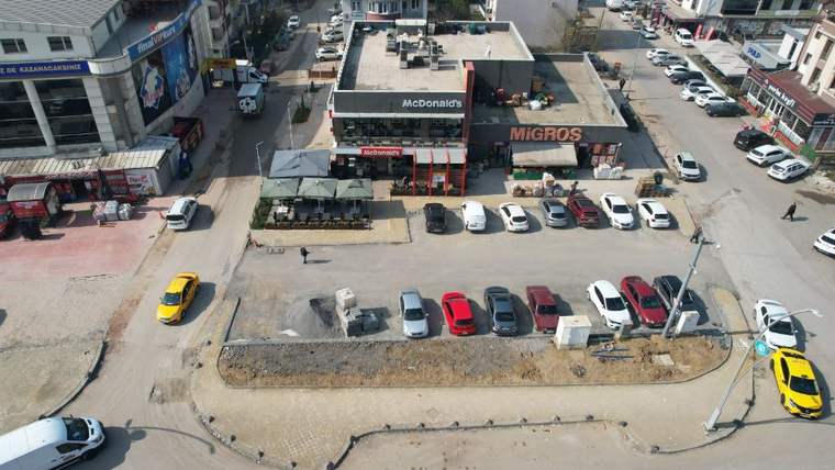 Havadan görünümde, çevresinde araçlar ve iş yerleri bulunan geniş bir otopark alanı görülüyor.