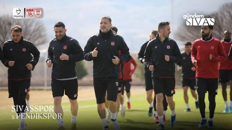 Sivasspor armalı formalar giyen futbolcular, açık havada çim sahada yan yana koşuyor.