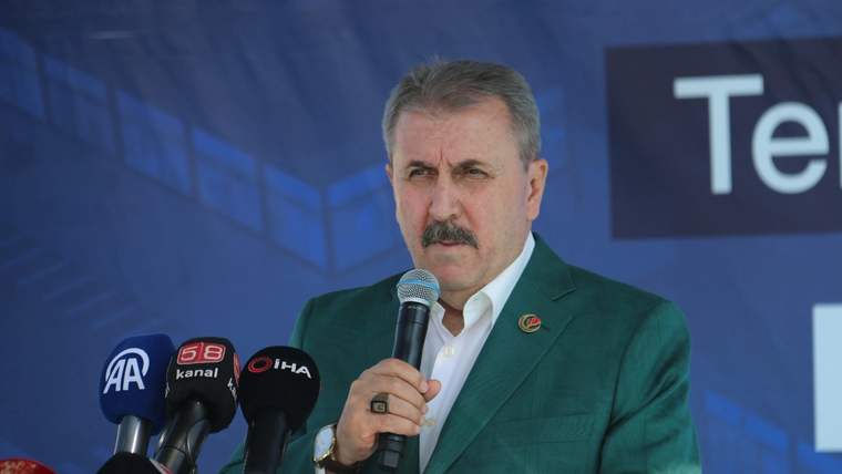 Bıyıklı bir kişi, elindeki mikrofonla konuşurken önündeki basın mikrofonları ve mavi arka plan görülüyor.