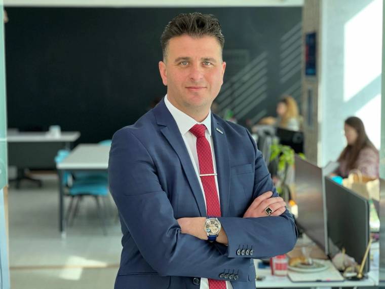 Koyu takım elbiseli bir kişi, kollarını bağlamış halde aydınlık bir ofis ortamında kameraya bakıyor.